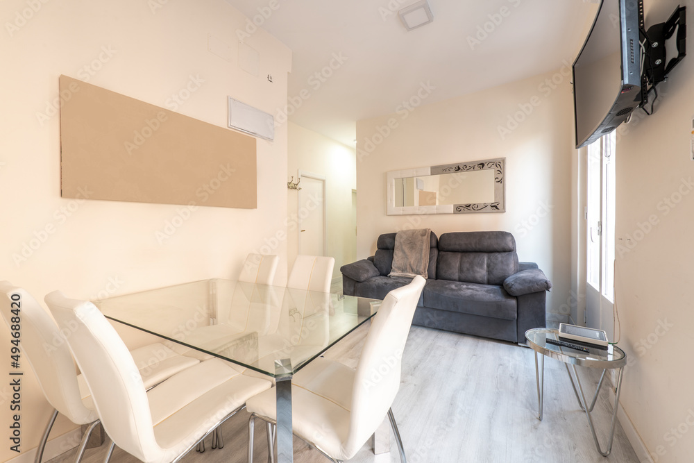 Glass dining table with white leather upholstered chairs in apartment with black leather sofa and wall mounted tv