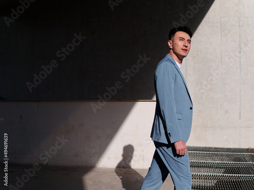 Confident businessman, portrait of handsome Chinese young man in light blue suit smiling and walking into sunlight, side view.