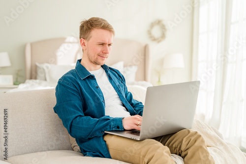 A young man in a blue shirt is working with a laptop while sitting on the couch at home in a cozy room. The concept of using the technology of the contact communication device business, IT technolog