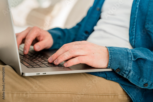 Hands a young man in a blue shirt using a laptop on his lap while sitting on the couch. Cozy conditions for remote work