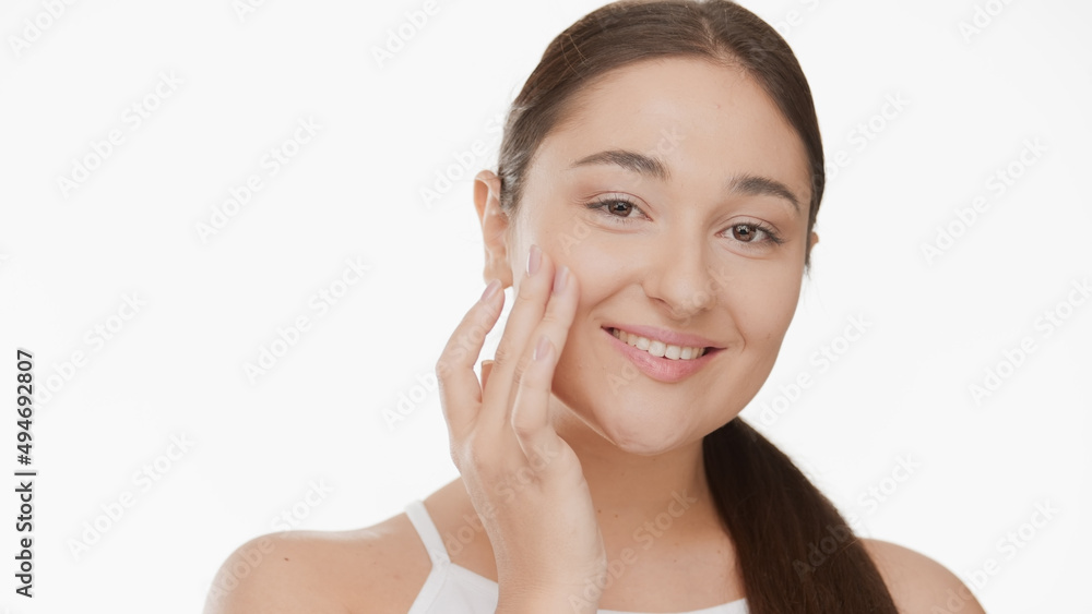 Attractive Caucasian young plus size woman with long brown hair in ponytail in white top touches her cheek smiling widely against white background | Skin nutrition concept Attractive Caucasian young plus size woman with long brown hair in ponytail in white top touches her cheek smiling widely against white background | Skin nutrition concept