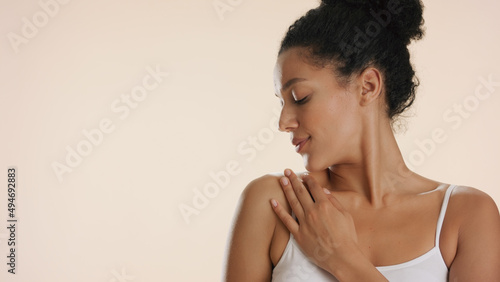 Young African American woman with curly hair in white bra strokes her clavicle and shoulder looking aside against beige background | Skin moisturizing concept