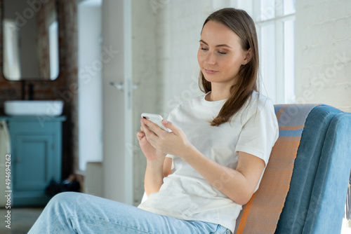 Smiling young woman is resting after work in the free space of the office or at home, sitting in an armchair, chatting with friends in a smartphone app