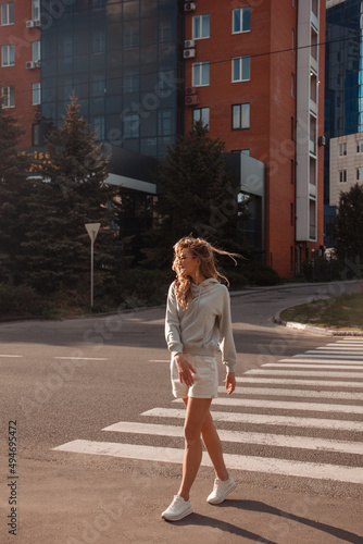 teen walk. young blonde curly girl in sports gray costume is standing in step near crosswalk and looking away in sunglasses on the street buildings background. sport lifestyle concept, free space