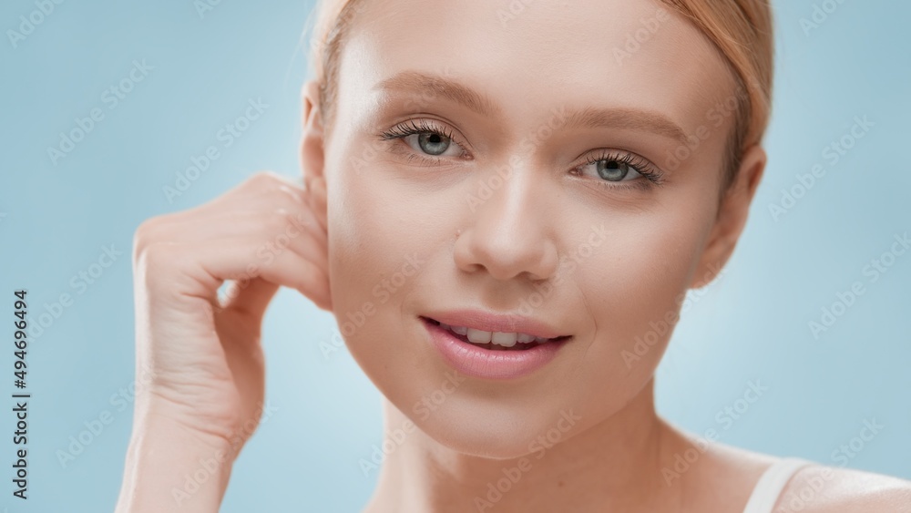 Young slim gorgeous European blonde female model touches jaw line and looks at camera against blue background | Skin moisturizing concept Young slim gorgeous European blonde female model touches jaw line and looks at camera against blue background | Skin moisturizing concept