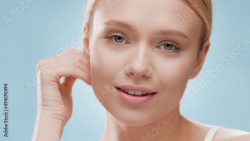 Young slim gorgeous European blonde female model touches jaw line and looks at camera against blue background | Skin moisturizing concept