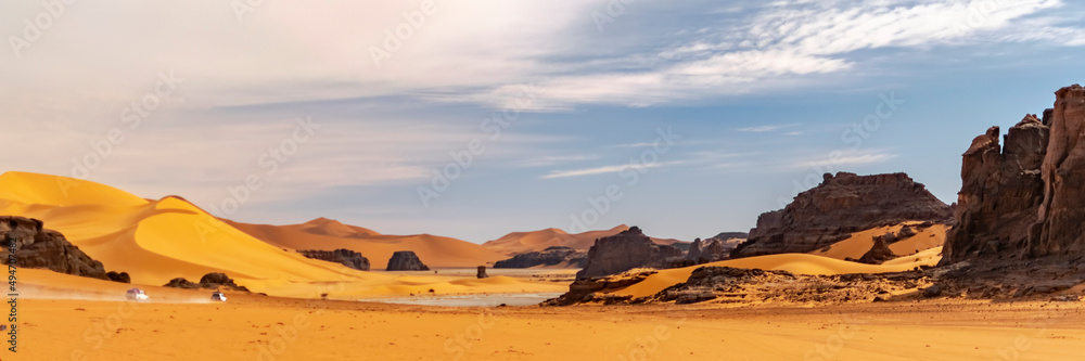 Panoramic view of Sahara Desert sand dune and rocky mountains in Tadrart Rouge, Djanet, Illizi. Far 4WD vehicles off-road speeding across the erg dusty road. Orange colored sandstones. Blue cloudy sky