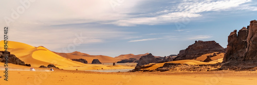 Panoramic view of Sahara Desert sand dune and rocky mountains in Tadrart Rouge  Djanet  Illizi. Far 4WD vehicles off-road speeding across the erg dusty road. Orange colored sandstones. Blue cloudy sky