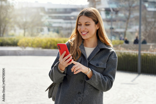 Medium shot of young smiling woman using cellphone in the street on sunny day photo