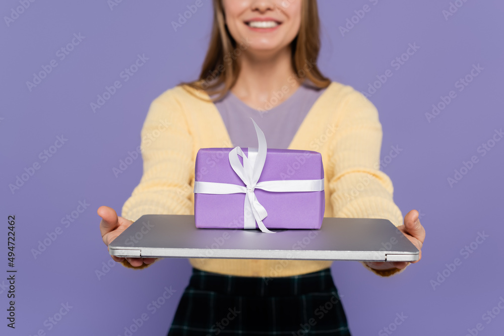 Obraz premium cropped view of happy young woman holding laptop with gift box isolated on purple.