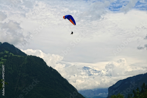 A man is flying on a paraglider over hills with trees, and in the background there are snow-capped mountains and the sky with clouds. Alpine landscape Interlaken, Switzerland