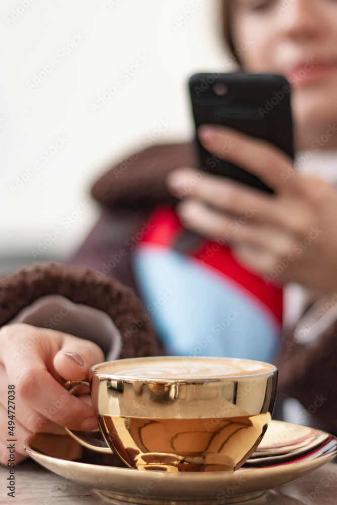 Young woman taking a photo of coffee with her smart phone. Girs face is not seen in photo. Focus is on the golden coffee cup. Girl is blur. Vertical photo. Young woman taking a photo of coffee with her smart phone. Girs face is not seen in photo. Focus is on the golden coffee cup. Girl is blur. Vertical photo.