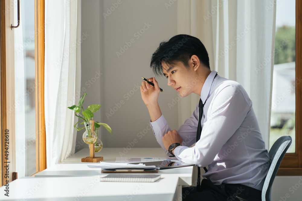 Portrait of a nice smiling grey asian man holding his pen, working at home on some project, he is sitting at a white table looking at the window in front of him. Focus on the man