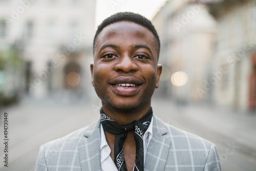 Portrait of handsome african american man in stylish suit and scarf smiling on camera while posing outdoors. Blur background of city street.