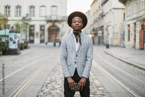 African male hipster in stylish suit and hat posing on city street with photo camera in hands. Happy young man enjoying free time on fresh air. #494730854