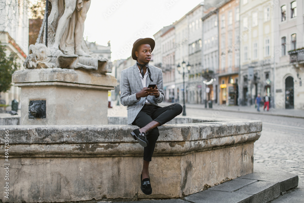 African american man in stylish suit and hat sitting on old fountain and holding modern smartphone. Handsome young guy surfing internet among city street. African american man in stylish suit and hat sitting on old fountain and holding modern smartphone. Handsome young guy surfing internet among city street.