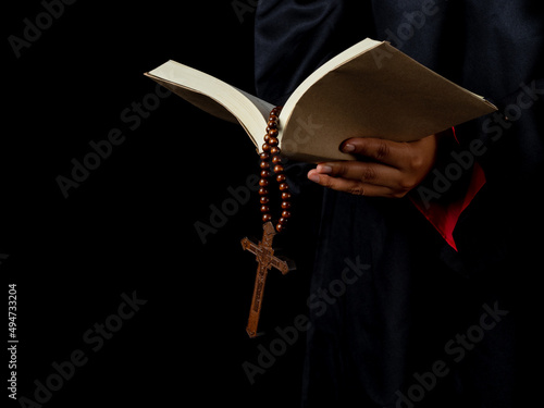 closed up hand holding Holy Bible with rosary beads and wooden carved crossed on black background.