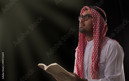 Mulim man in traditional clothing holding holy book while looking up praying with faith on black background. photo