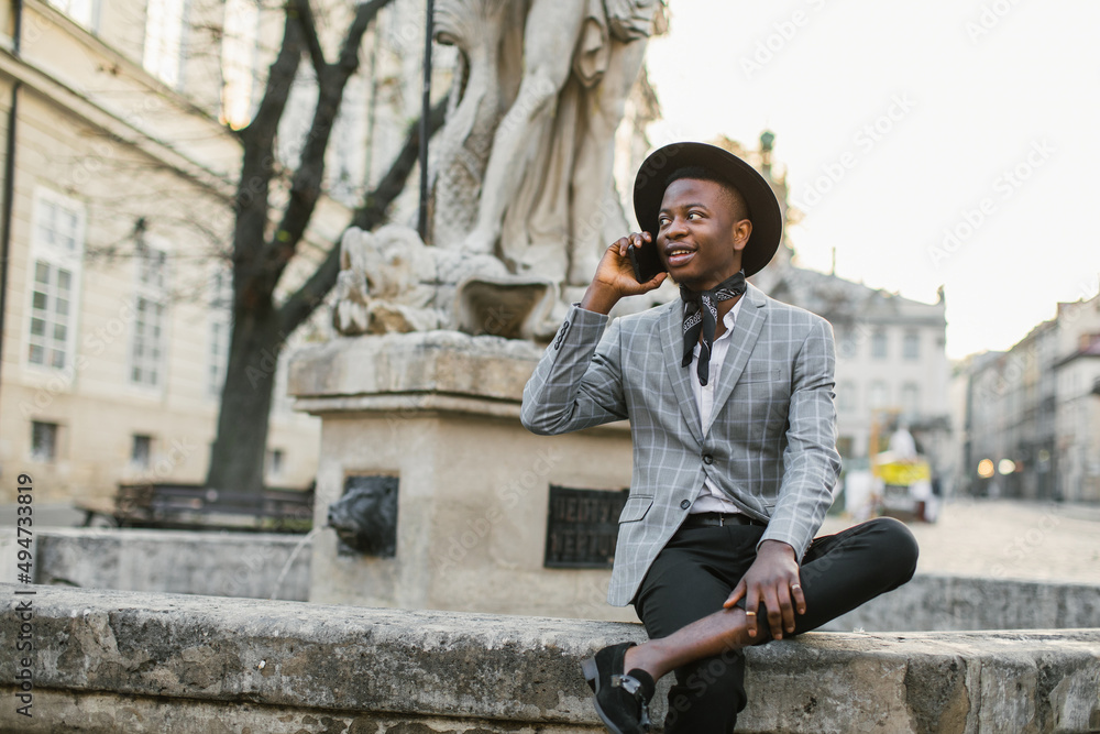Smiling african man in stylish wear having mobile conversation while resting outdoors. Young guy sitting on old fountain among city street.