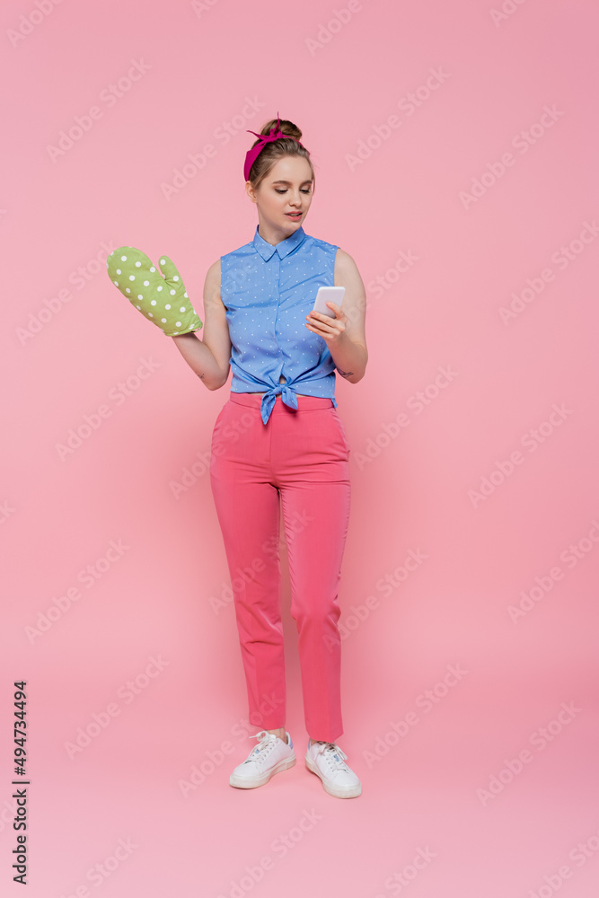 full length of young woman with green baking glove holding smartphone isolated on pink. full length of young woman with green baking glove holding smartphone isolated on pink.