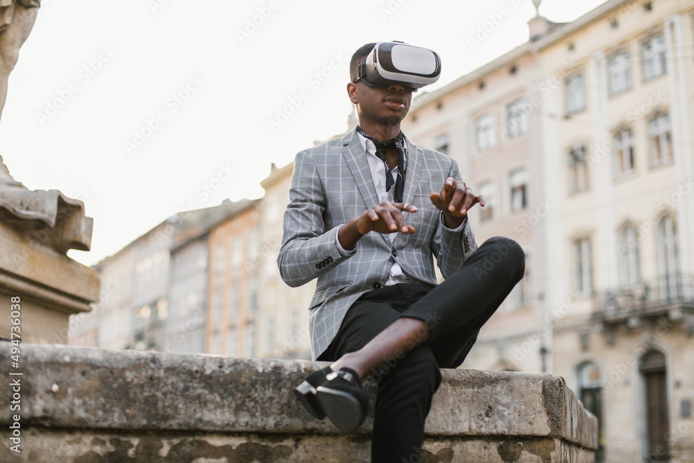 Successful african businessman in stylish wear using VR headset while sitting on city street. Young male person gesturing with hands in air while touching virtual screen. Successful african businessman in stylish wear using VR headset while sitting on city street. Young male person gesturing with hands in air while touching virtual screen.