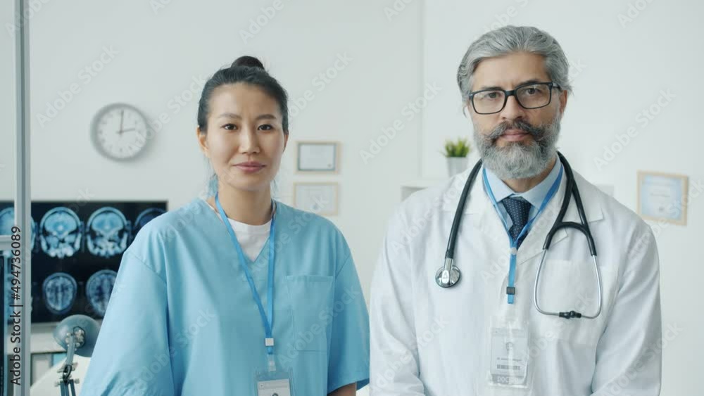 Portrait of male physician in white gown and female nurse in scrubs standing in clinic office and looking at camera. People and hospital concept. Portrait of male physician in white gown and female nurse in scrubs standing in clinic office and looking at camera. People and hospital concept.