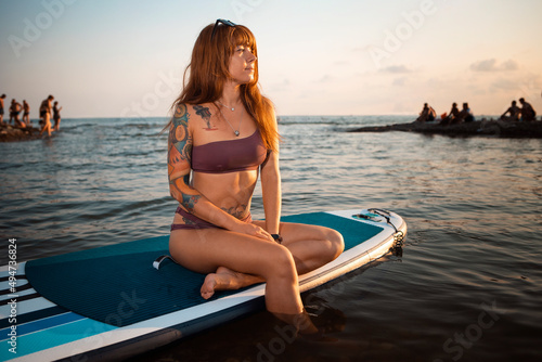 A tanned young woman with a tattoo poses sitting on a sup board. The concept of sup boarding and surfing © _KUBE_