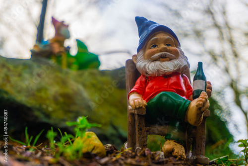 A yard gnome relaxes in his rocking chair with a garden gnome in the backdrop, riding a lizard looking on. photo