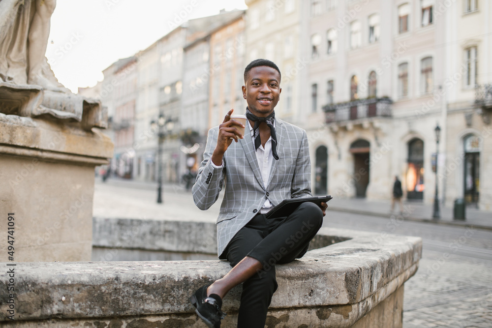 African american man in stylish suit sitting on old fountain with cup of coffee. Young male hipster enjoying hot beverage while resting on city street.