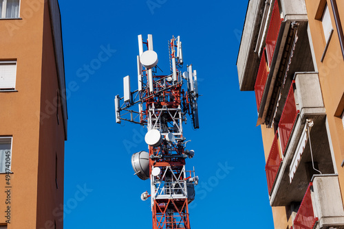 Metal tower with telecommunication antennas and parables colored red and white between residential buildings on blu sky, high speed 5G network and Electromagnetic pollution concept