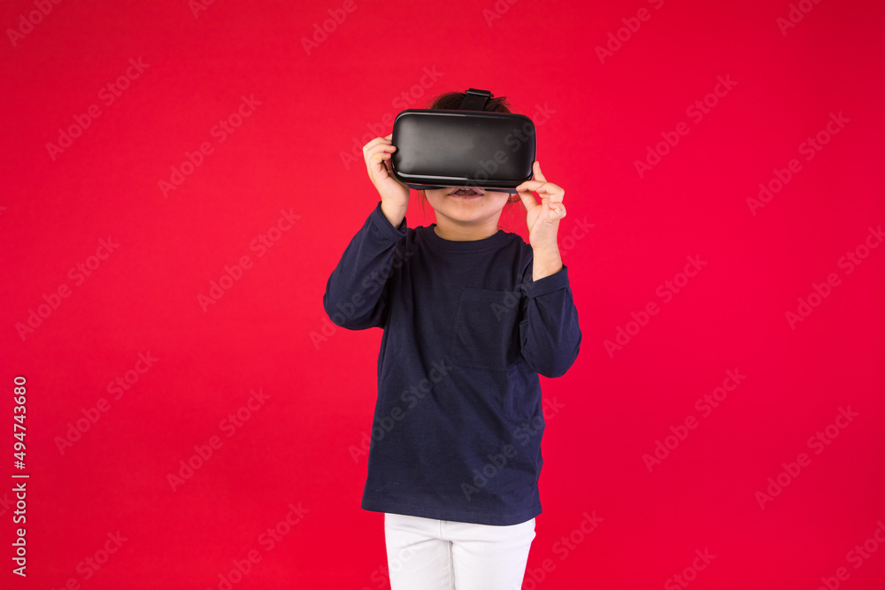 Front view of little girl wearing virtual reality glasses amazed and touching with her hands virtually, on red background. Virtual, augmented reality, technology, games and childhood concept. Front view of little girl wearing virtual reality glasses amazed and touching with her hands virtually, on red background. Virtual, augmented reality, technology, games and childhood concept.