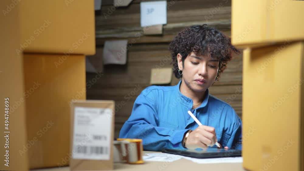 Selective focus of Asian female factory worker in a blue uniform standing among the cardboard piles, scanning barcode on a blurred cardboard boxes using a barcode scanner and take notes on a tablet. Selective focus of Asian female factory worker in a blue uniform standing among the cardboard piles, scanning barcode on a blurred cardboard boxes using a barcode scanner and take notes on a tablet.