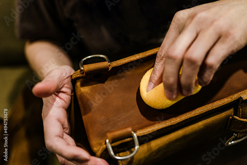 Professional leather care service concept. Closeup hand with yellow soft sponge cleaning surface of the vintage brown bag, depth of field. Polish waxing protection from dust and dirt.