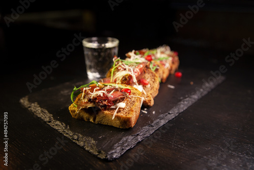 Bruschetta with meat  herbs  and sauce. Restaurant serving on a blackboard on a dark background. A glass of alcohol on the background.