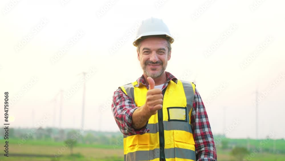 Happy caucasian senior man engineer standing and thumb up to camera while smiling in front of wind turbine farm at behind. Happiness and proud in occupation of alternative power industrial worker. Happy caucasian senior man engineer standing and thumb up to camera while smiling in front of wind turbine farm at behind. Happiness and proud in occupation of alternative power industrial worker.