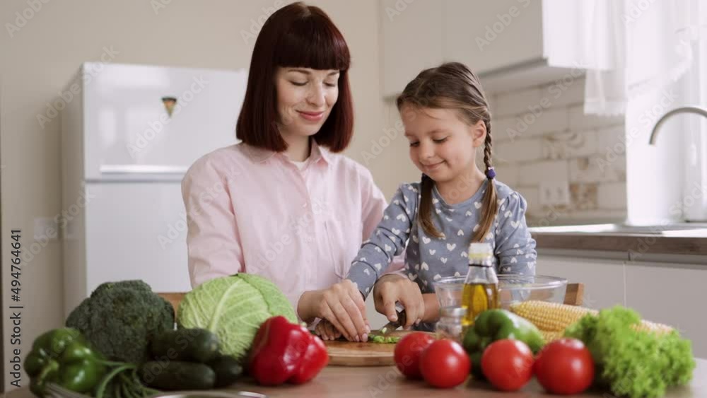 Cute little girl and her beautiful mom in casual clothes are cutting onion and smiling while cooking in kitchen at home. Healthy food at home. Happy family in the kitchen.