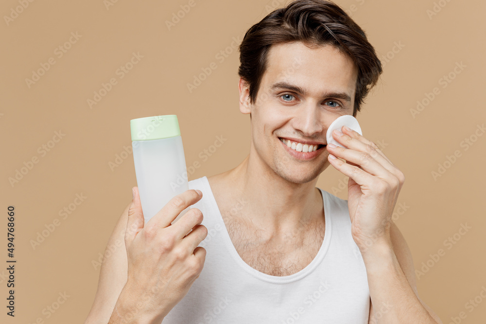 Young smiling man 20s perfect skin in undershirt hold bottle tonic apply moisturizing lotion with cotton pad isolated on pastel pastel beige background Skin care healthcare cosmetic procedures concept Young smiling man 20s perfect skin in undershirt hold bottle tonic apply moisturizing lotion with cotton pad isolated on pastel pastel beige background Skin care healthcare cosmetic procedures concept