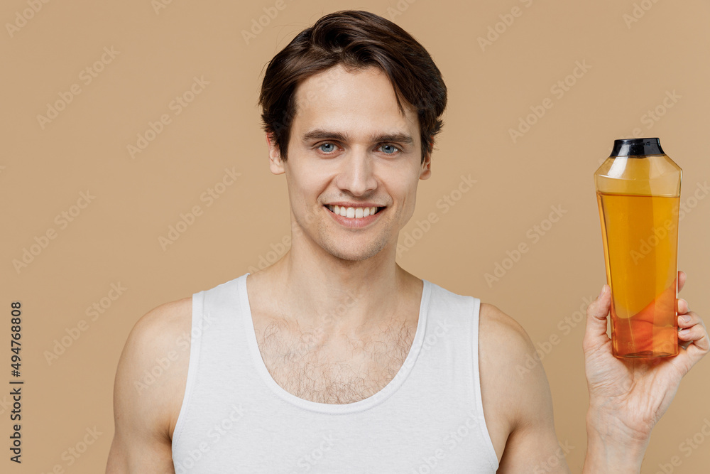 Attractive smiling happy young man 20s perfect skin in undershirt hold bottle of shampoo isolated on pastel pastel beige background studio portrait. Skin care healthcare cosmetic procedures concept. Attractive smiling happy young man 20s perfect skin in undershirt hold bottle of shampoo isolated on pastel pastel beige background studio portrait. Skin care healthcare cosmetic procedures concept.