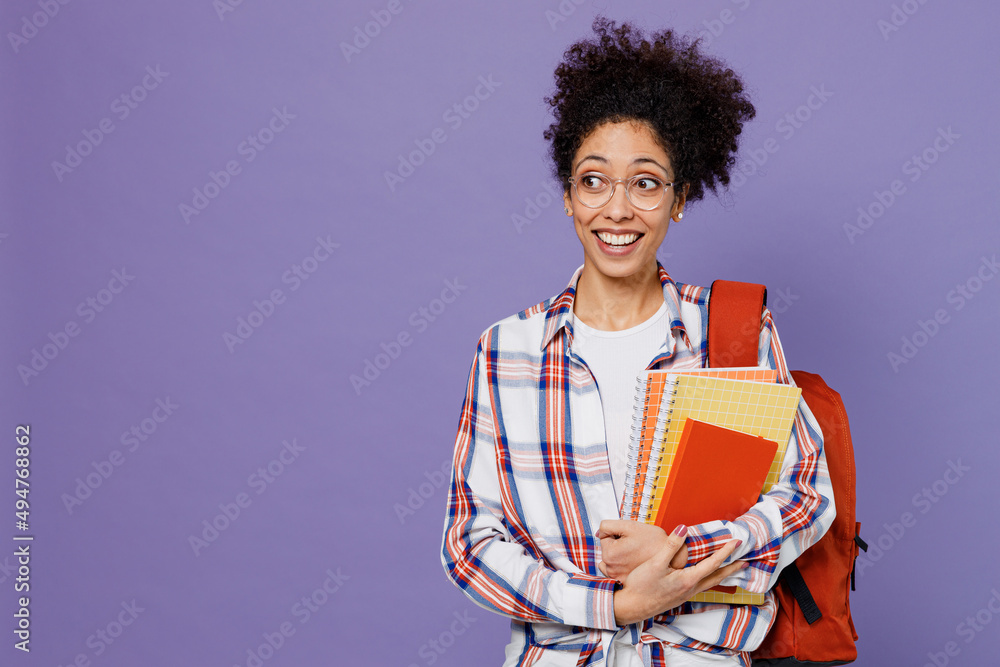 Young surprised girl woman of African American ethnicity teen student in shirt backpack hold books look aside isolated on plain purple background. Education in high school university college concept. Young surprised girl woman of African American ethnicity teen student in shirt backpack hold books look aside isolated on plain purple background. Education in high school university college concept.