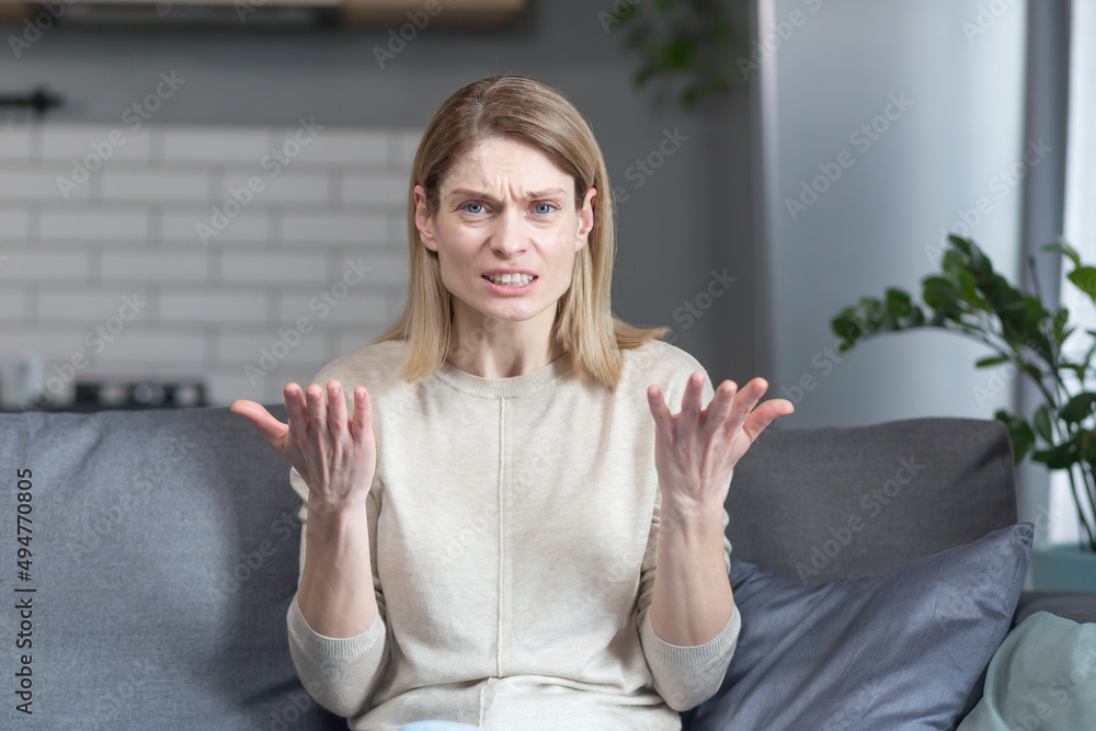 angry and frustrated woman talking on video call looking at webcam, angrily gesturing with hands while sitting on sofa at home angry and frustrated woman talking on video call looking at webcam, angrily gesturing with hands while sitting on sofa at home
