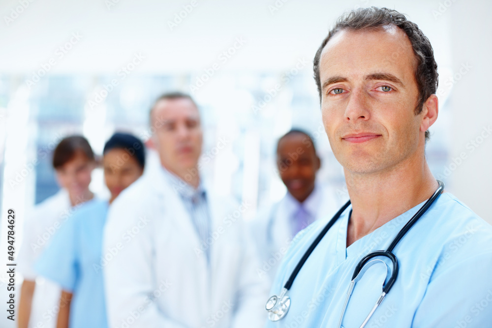Confident resident in front of team. Portrait of serious resident with medical team in background. Confident resident in front of team. Portrait of serious resident with medical team in background.
