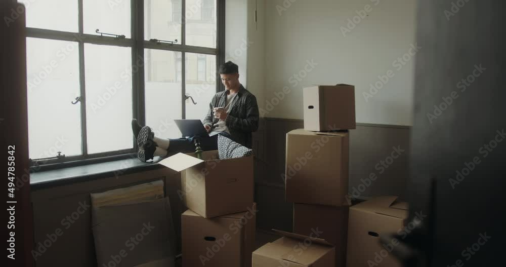 Asian Adult Male using Laptop in New Home Planning Surrounded by Boxes Asian Adult Male using Laptop in New Home Planning Surrounded by Boxes