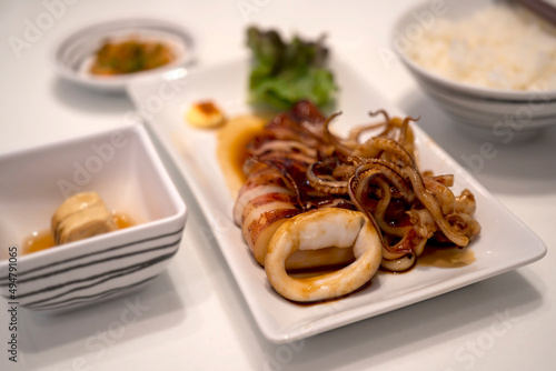 A Japanese set of Grilled squid with soy sauce served in a white plate with green vegetable.