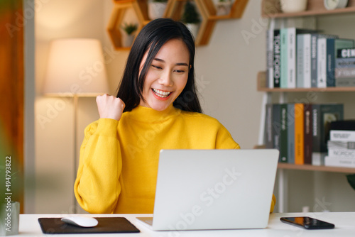 Young Asian woman using laptop showing winner gesture, glad, excite at home