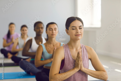 Portrait of a relaxed athletic woman enjoying meditation with closed eyes at a yoga class at a sports club. Woman sits in a lotus position with folded palms in a group yoga class. Blurred background.