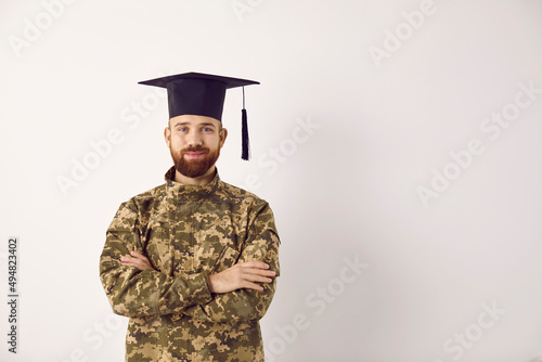 Bild auf Leinwand Portrait of happy young military student wearing soldier uniform and square academic graduate cap standing against copy space wall background, looking at camera and smiling