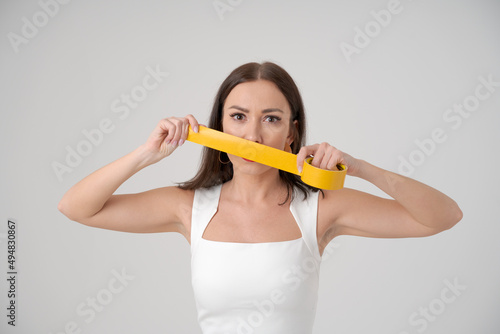 Frightened woman holding tape in front her face covering her mouth and lips. Concept prohibition statements. Caucasian woman is afraid to say her opinion in a white stylish dress on light background