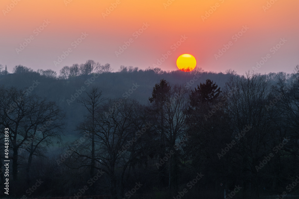 A spring sunset over het rolling hill landscape in the south of the Netherlands with a view on the green meadows the forest and in the background the sun going down, to cover the landscape in the dark