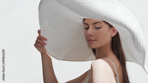 Young attractive European woman with long dark hair in a big white hat and white bikini holds a brim of her hat and looks aside against white background | Spf cream concept