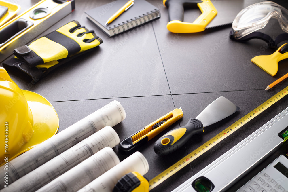 Business contraction theme. Tool kit of the contractor: yellow hardhat, libella, hand saw. Plans and notebook on the gray tiles. Business contraction theme. Tool kit of the contractor: yellow hardhat, libella, hand saw. Plans and notebook on the gray tiles.